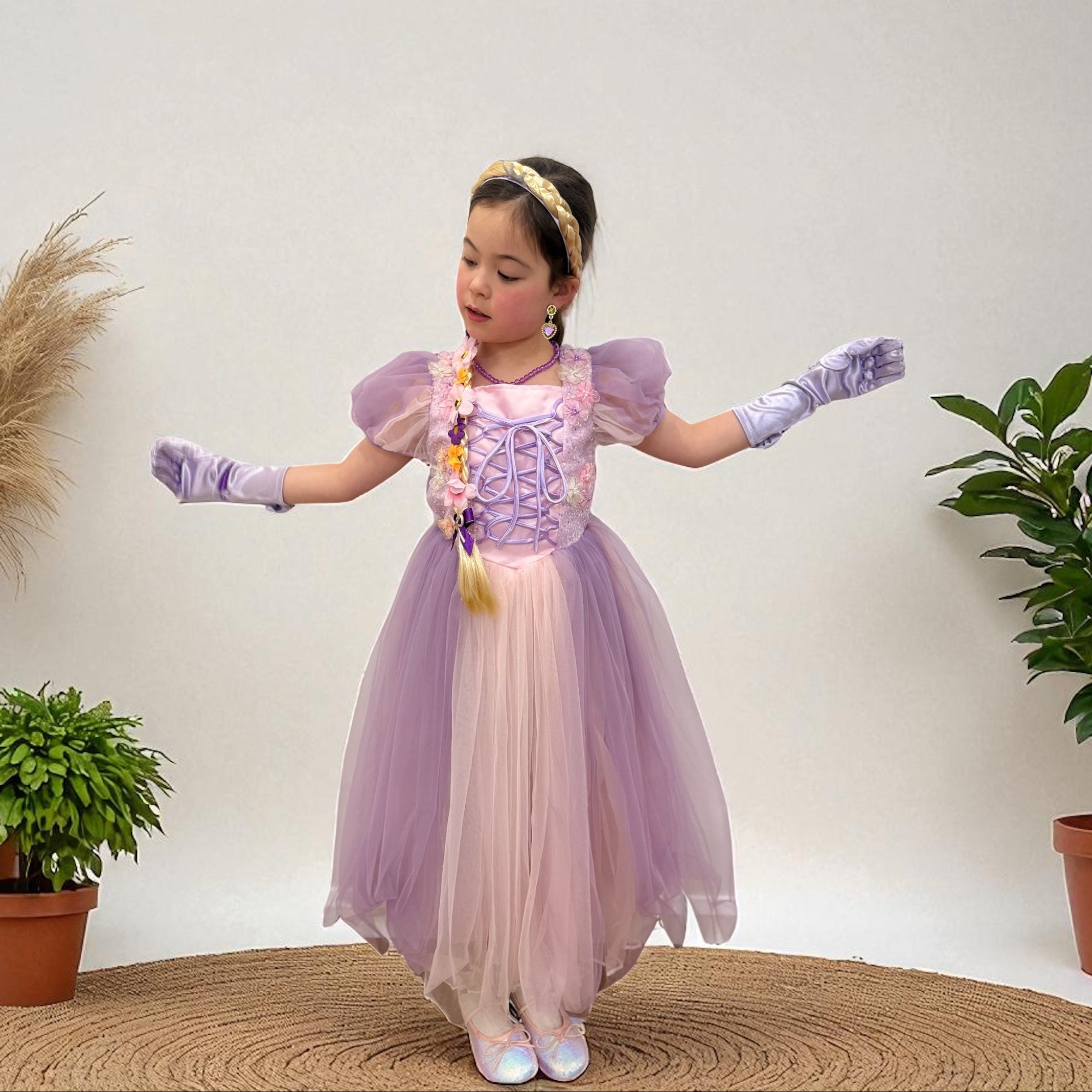 Little girl wearing a lavender princess dress with puff sleeves, matching satin gloves, and a floral braid accessory against a neutral studio background.